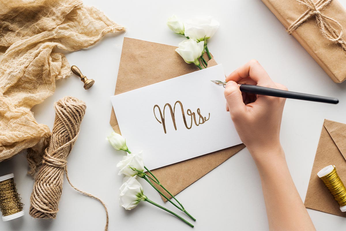 White hand holding a pen addressing an envelope to “Mrs.” on a white table with brown twine, a cloth, some gold thread, and white carnations.