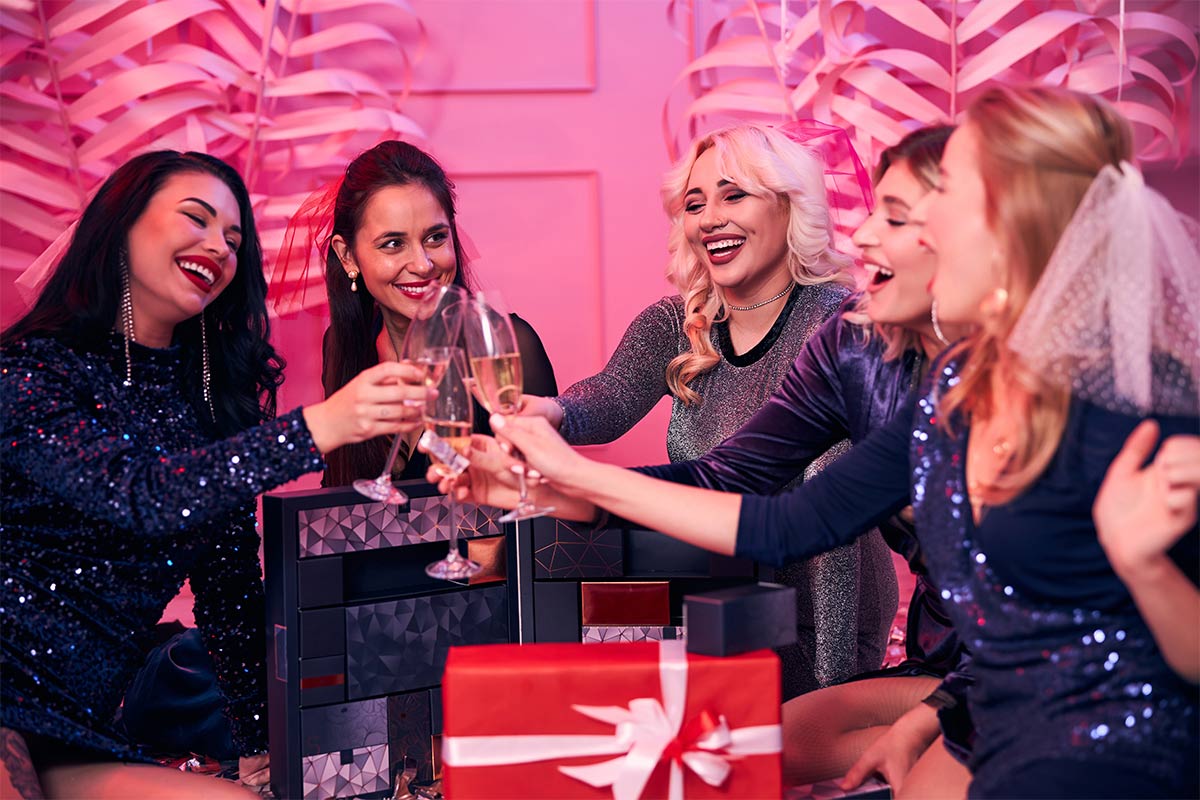 Five white woman in their 30s wearing sequin and sparkly dresses smile and cheers with champagne over gift boxes while sitting in a pink room 