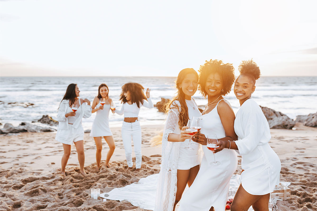 Three multiracial women in their 30s wearing all white stand and smile for a photo with red drinks on the beach during sunset as three other women wearing white dance in the sand in the background 