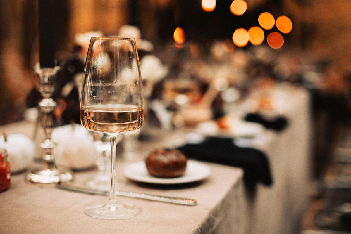 fancy glass of white wine sits on an elegant dinner table that contains a blurred spread of napkins, candles, and plates