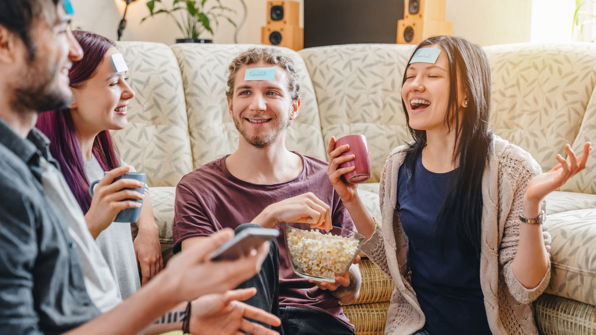 Four white young people sit on a couch playing a guessing game with sticky notes on their foreheads, holding drinks and popcorn, laughing and chatting.