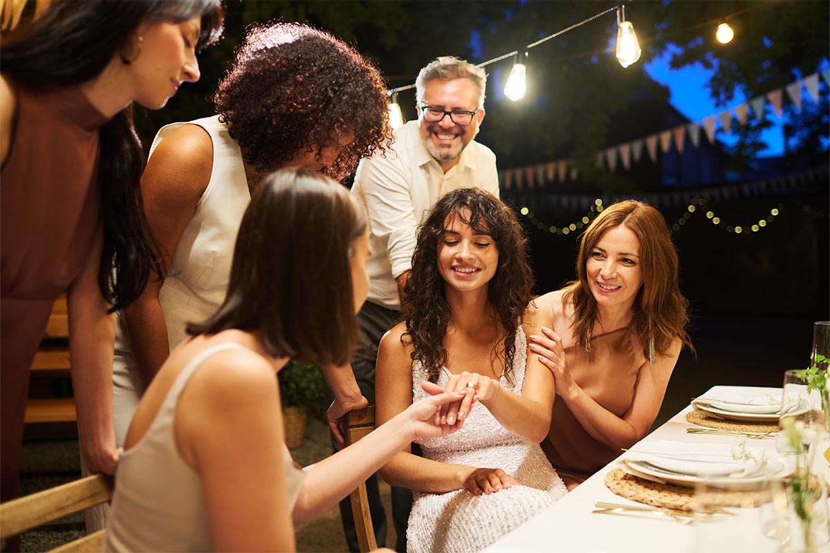  Group of multiracial friends in their 30s and 40s sit and stand outside at a table during night time as they admire a ring on a 30 something white woman’s finger