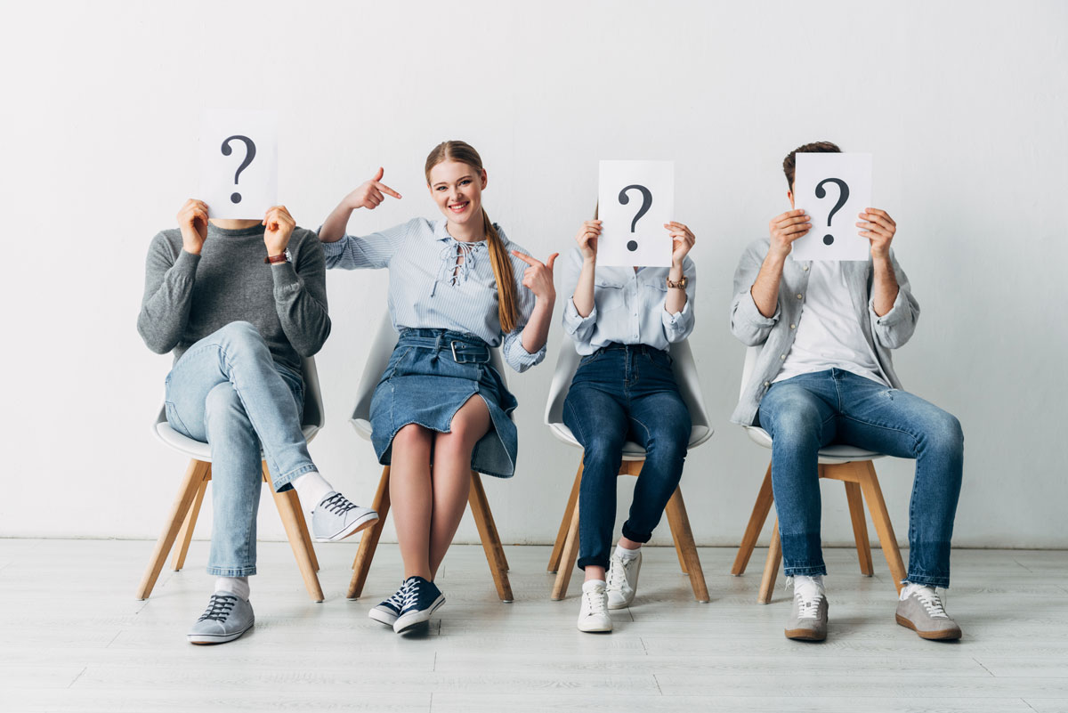 A white 20-year-old woman sitting in between three people holding question marks in front of their faces. 