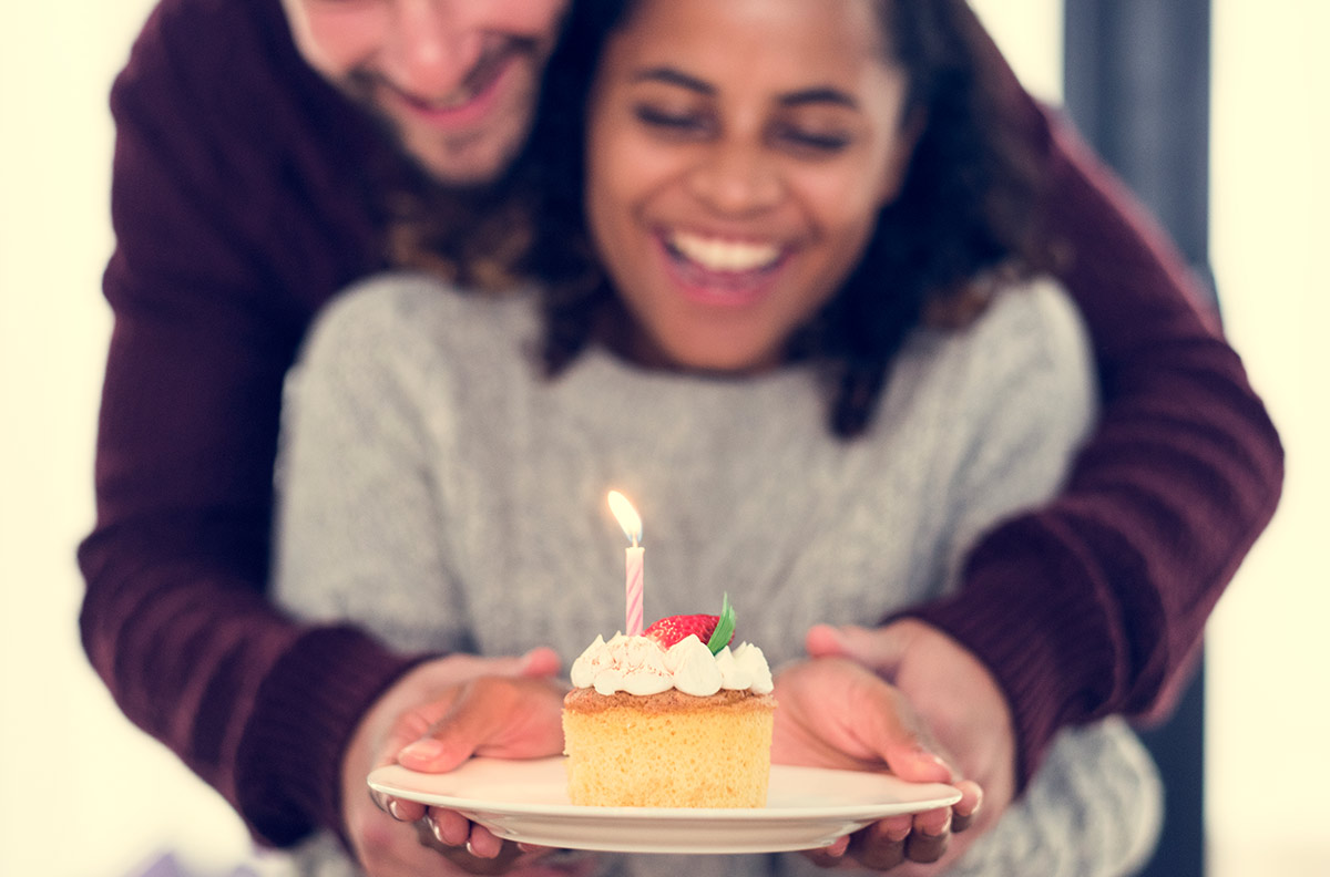 30 something white man hands 30 something black female partner a cupcake with a lighted on candle on top while embracing her from behind