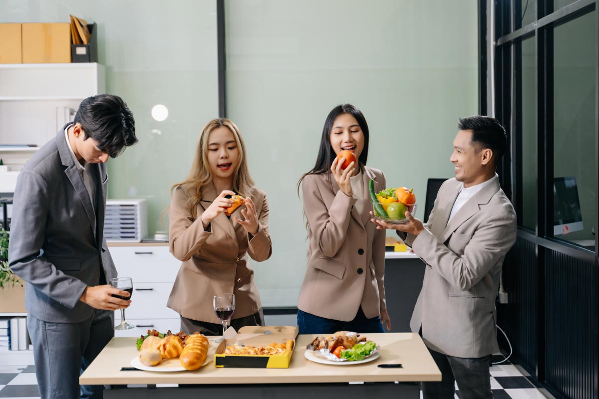 A group of 4 Asian coworkers wearing suits, eating a snack board and standing around the snack board.