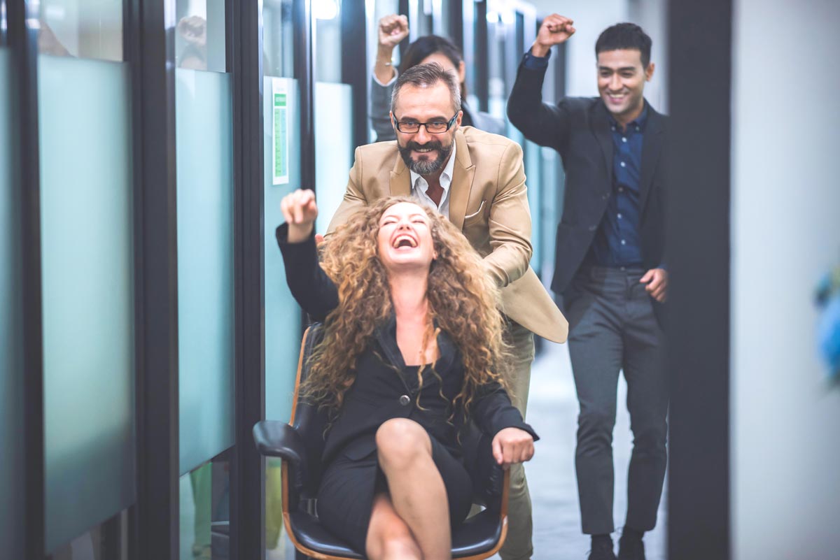 A group of 4 cheerful white office employees celebrate together in a hallway, a woman with curly hair and a joyful expression sits in a rolling chair, as a man in a tan blazer pushes her from behind.