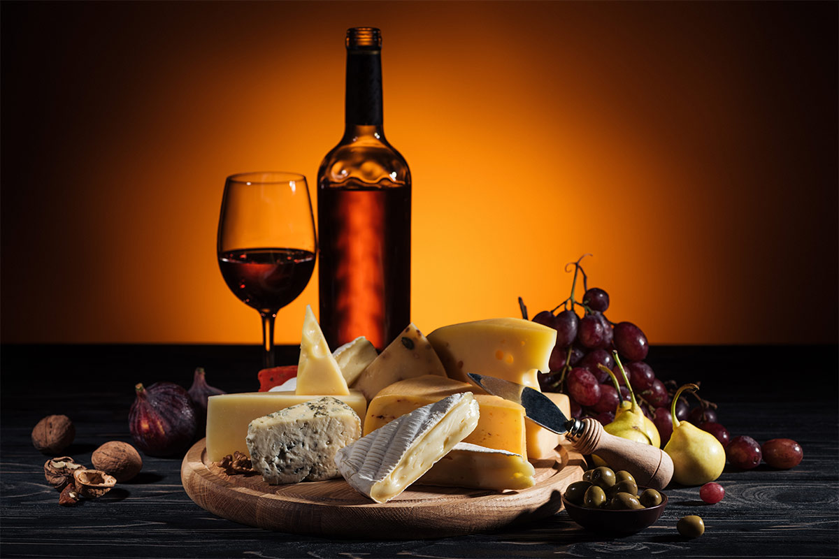 cheese, grapes, nuts, olives, and other foods sit on and around a wooden plate on a black surface while a bottle and glass of wine shine due to an orange light in the background