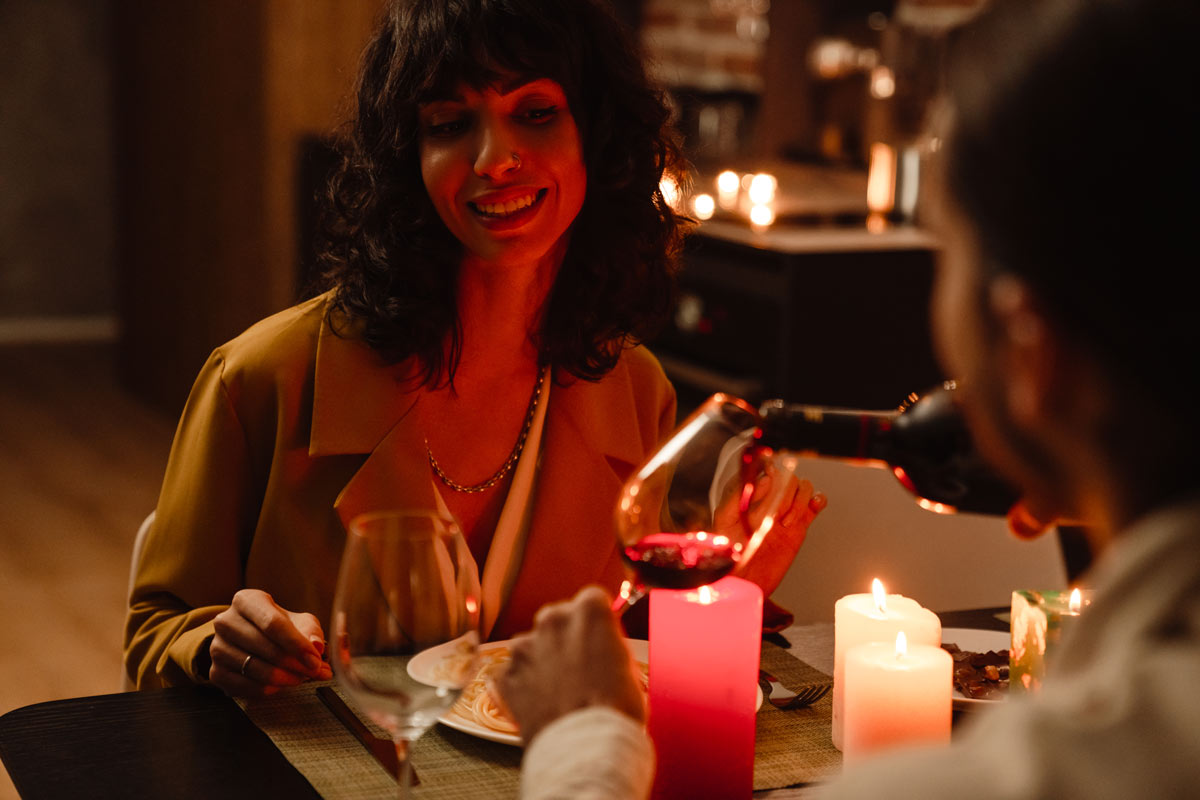 A man pours a glass of red wine for a white 30-something woman at a candlelit dinner.