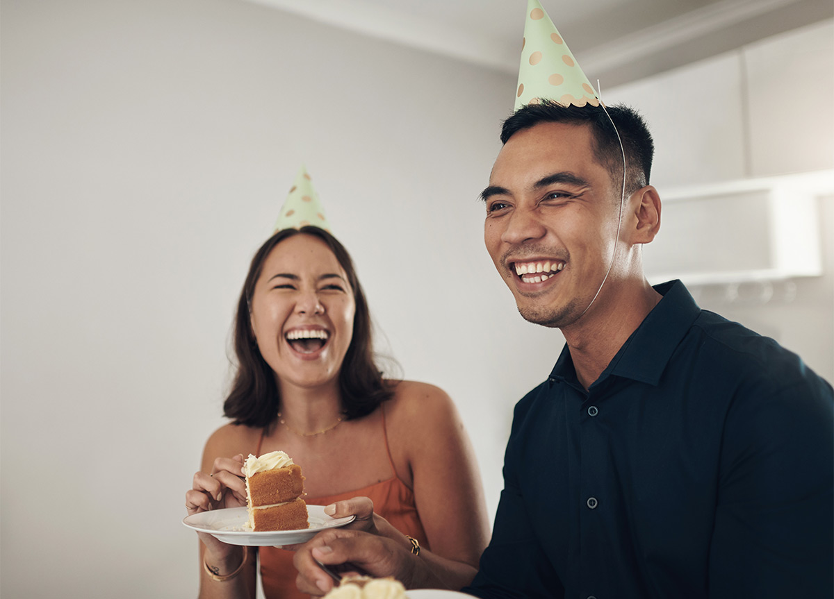 30-something female and male with birthday hats laugh and eat cake indoors