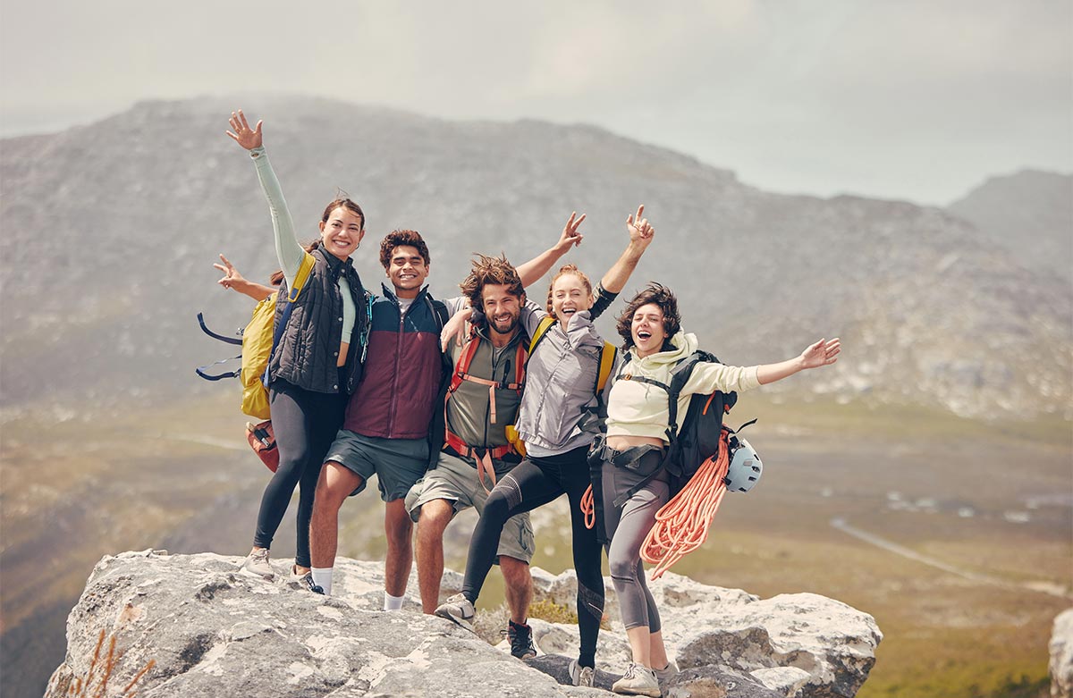 Group of multiracial friends in their 30s with hiking gear stand and pose with arms in the air on a high rock in the mountains during the day