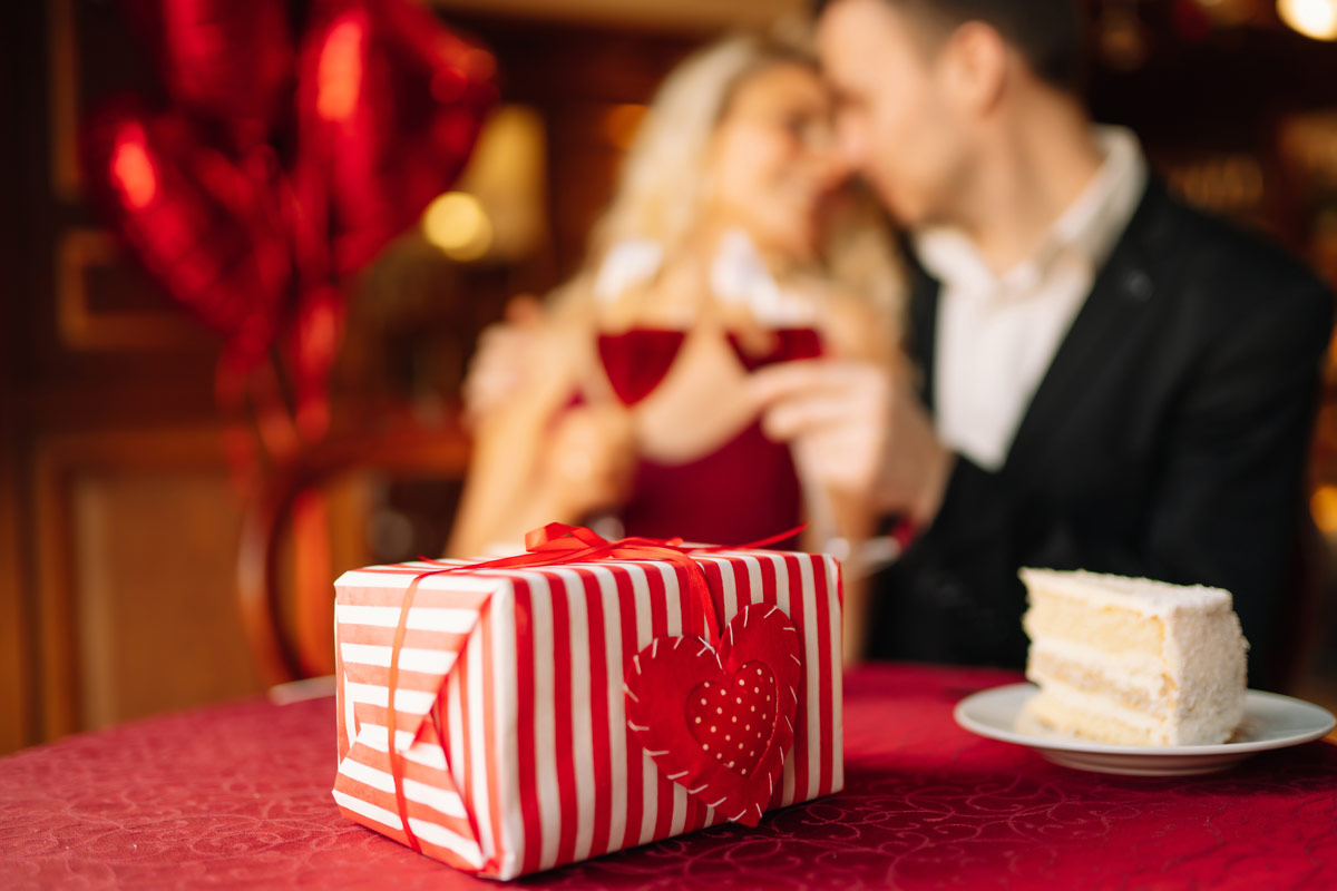 a 20 something white couple in formal attire embrace while drinking wine together in a room with heart balloons and a present and piece of cake sitting on top of a table with a red tablecloth.