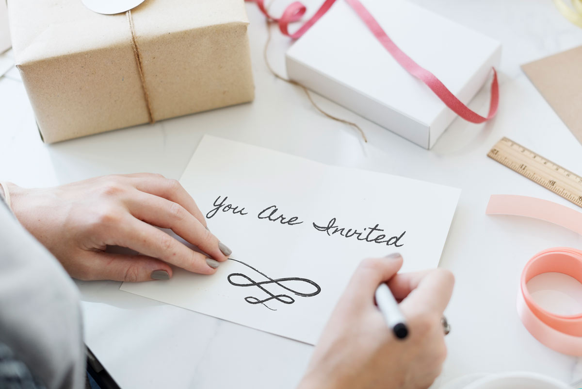 A woman writing on a piece of white paper “you are invited” to show that she has made her guest wedding selections and is getting ready to formally invite them to the wedding.