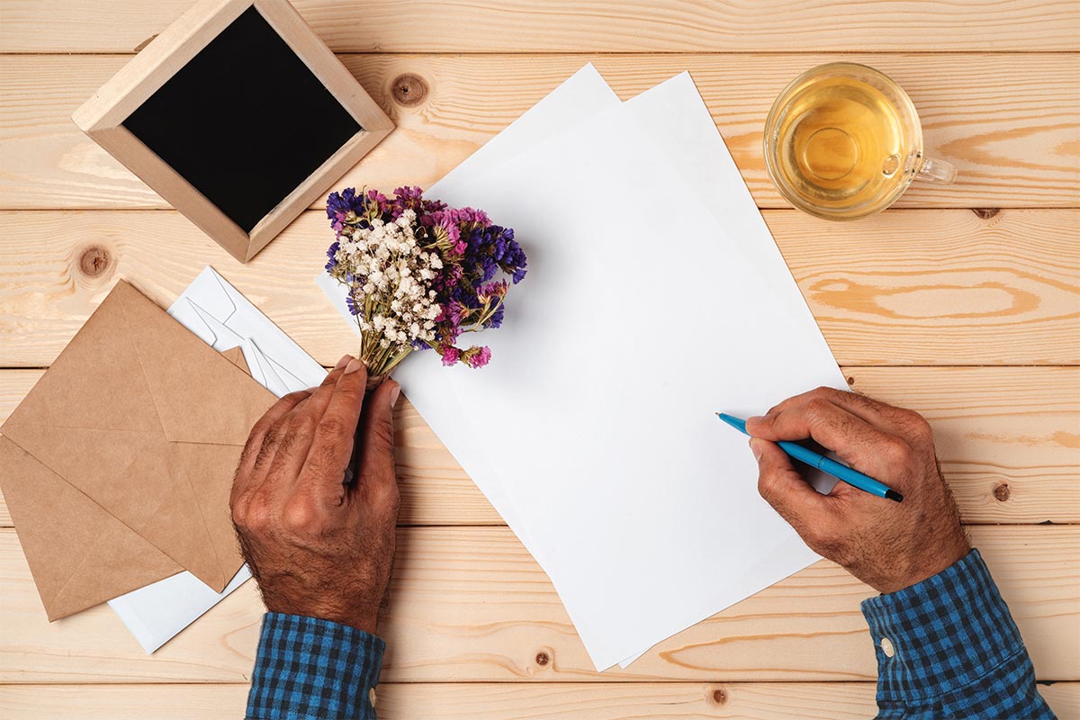 Older Hispanic male hands with pink, white, and purple small flowers in one hand and a blue pen in the other about to write a letter with a picture frame and tea on the wooden desk.