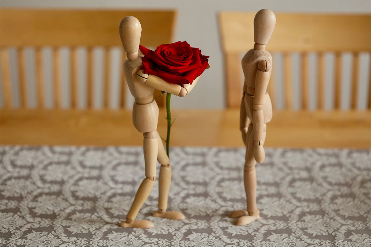 2 wooden artist figures on a table with one giving a red rose to the other 