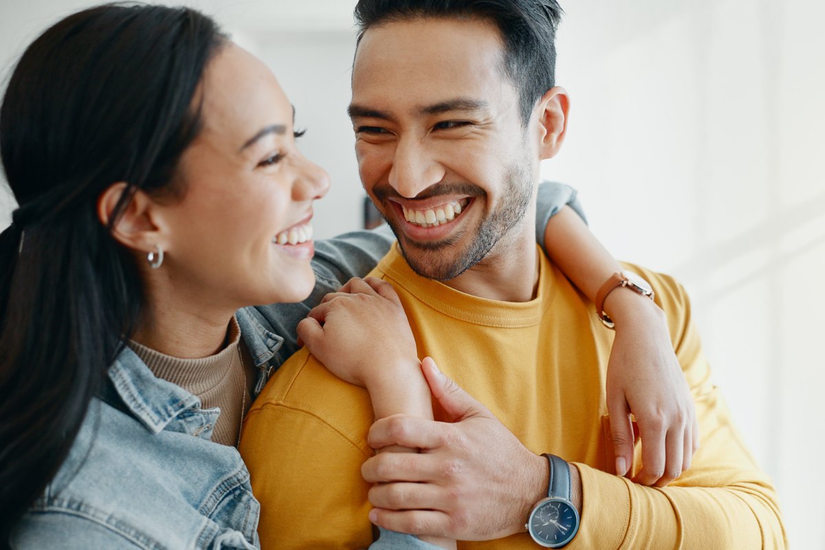 mixed 30-something woman hugging asian 30-something man from behind as he’s looking at her smiling inside