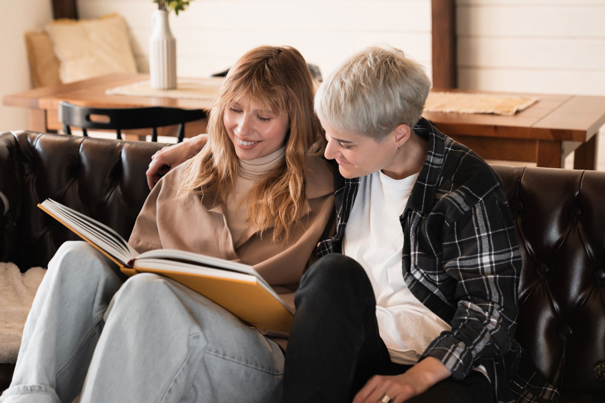 Two middle aged white women looking at a book together smiling sitting on a couch 