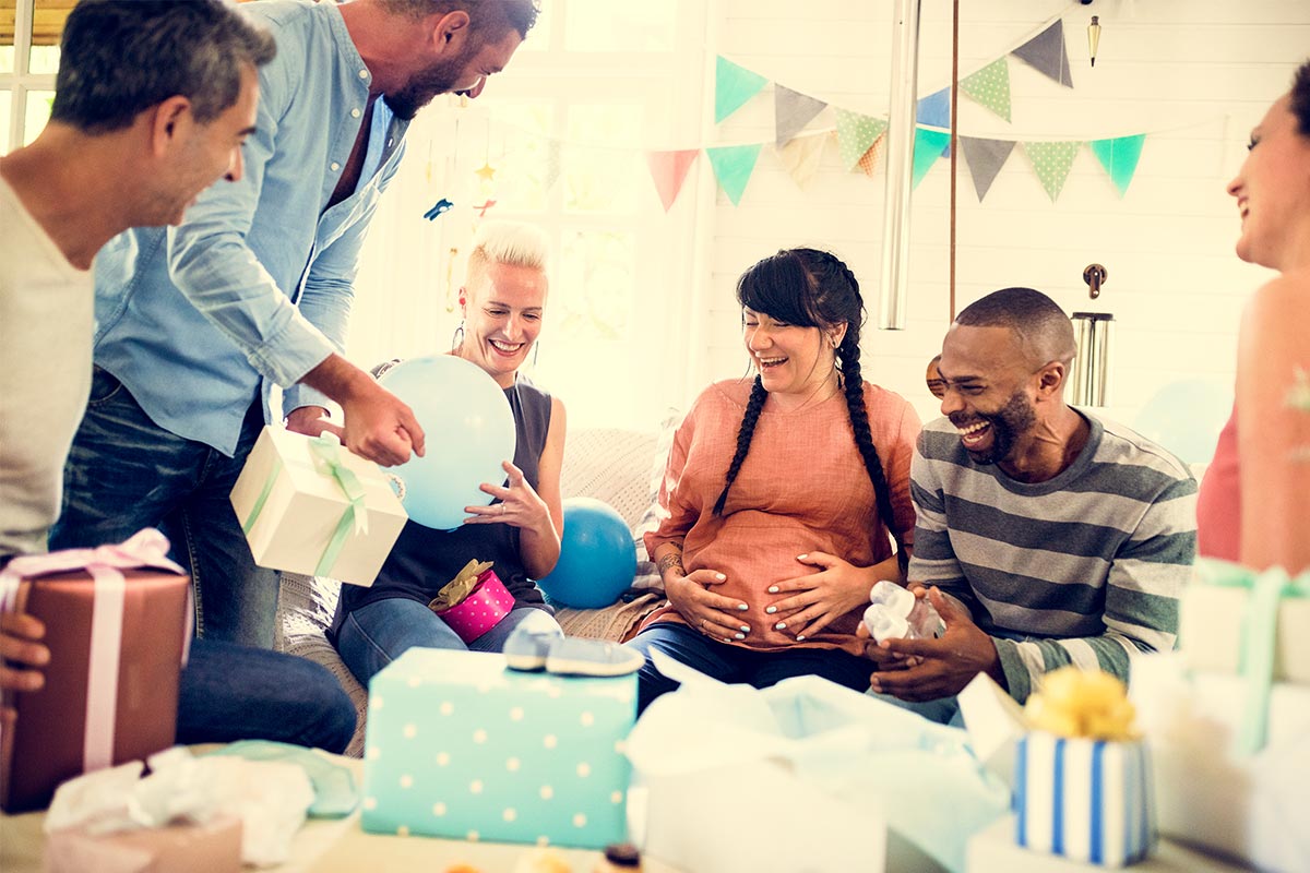 A pregnant white woman in her 30s receives wrapped presents at a party as she sits on couch surrounded by multiracial friends in their 30s and 40s