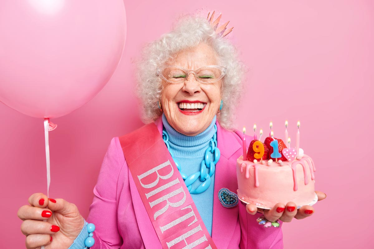 White 91 year old woman in pink birthday sash and crown smiles big as she holds a pink strawberry birthday cake with lit candles and a large pink balloon