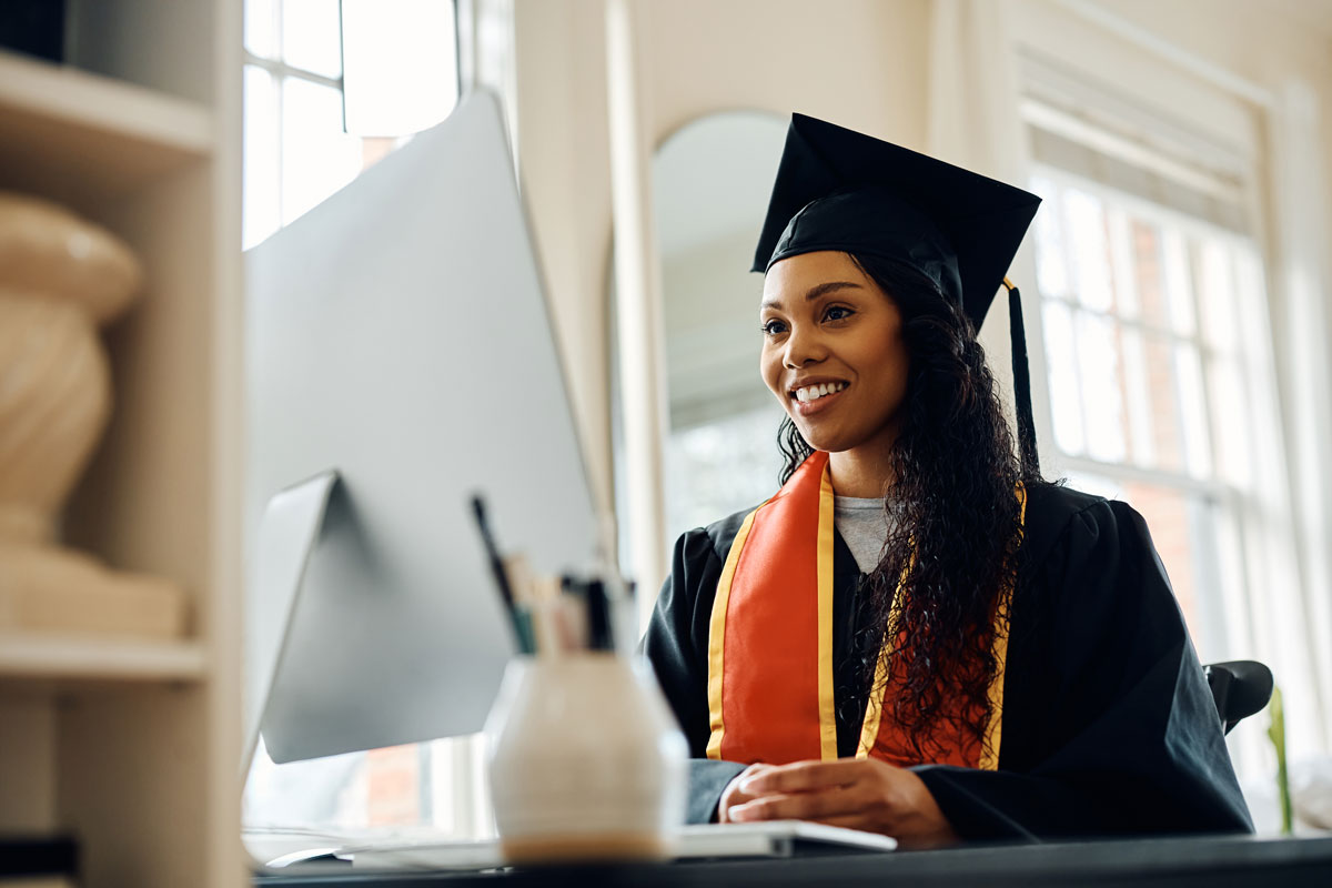 Smiling 23-year-old black woman wearing a graduation cap and gown, at a desk with a computer, celebrating her graduation virtually with family and friends online.