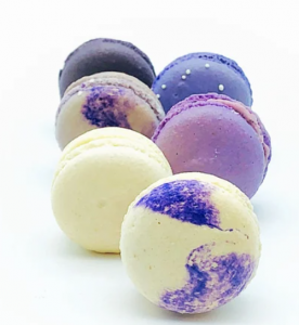 Different colored macaron cookies. Colors include purple, yellow, blue with sprinkles, black, and two cookies with a mixture of yellow and purple.