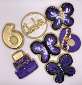 Custom purple and gold colored cookies. Types of cookies include three butterfly cookies, two cookies with numbers on them, a cookie with a three-tiered cake on it, and a cookie with the name "Charlotte" written across in gold ink.